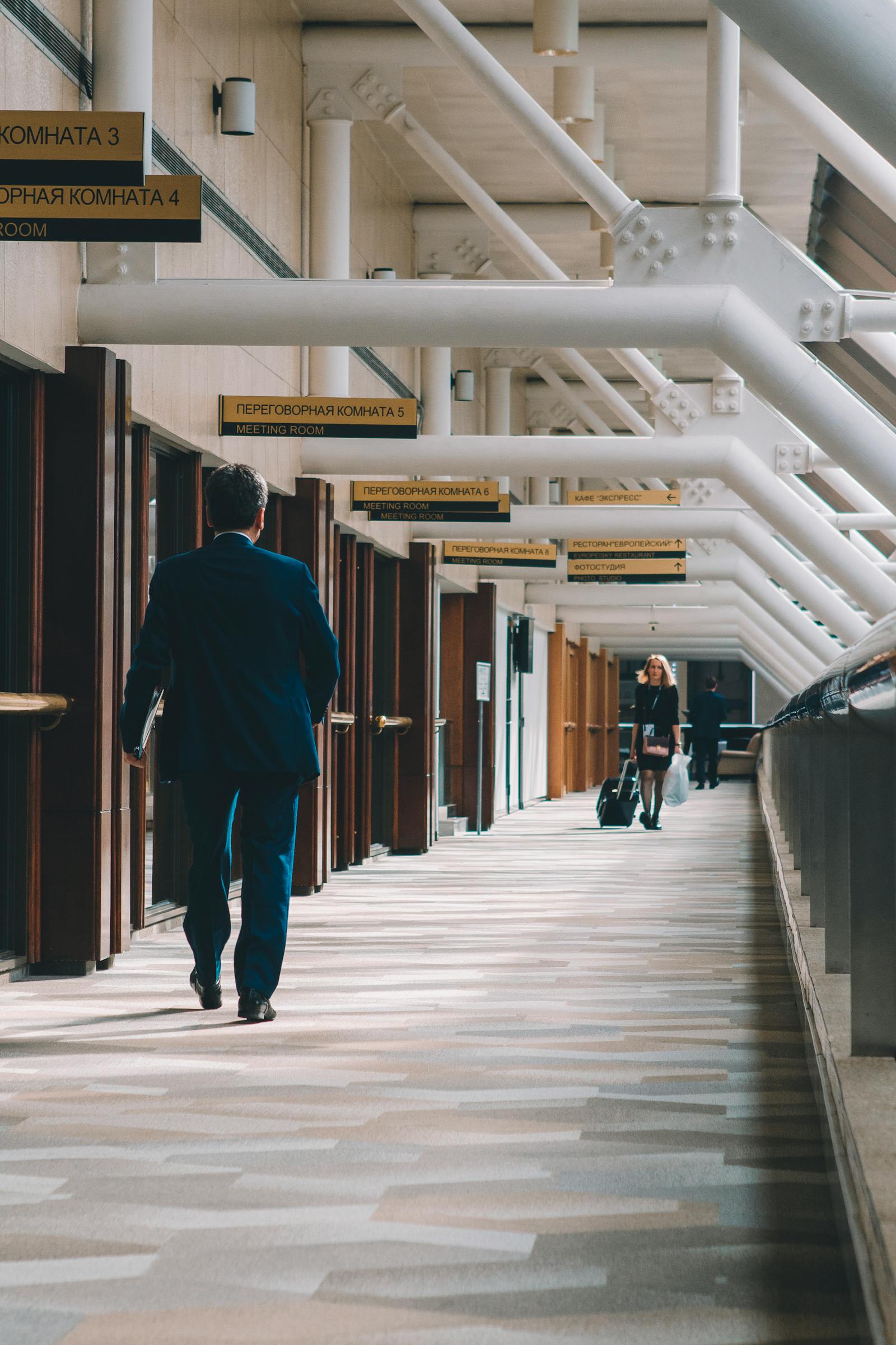 A man in a navy suit walks down a long, sign-lined corridor; a colleague with a wheeled bag in the distance.
