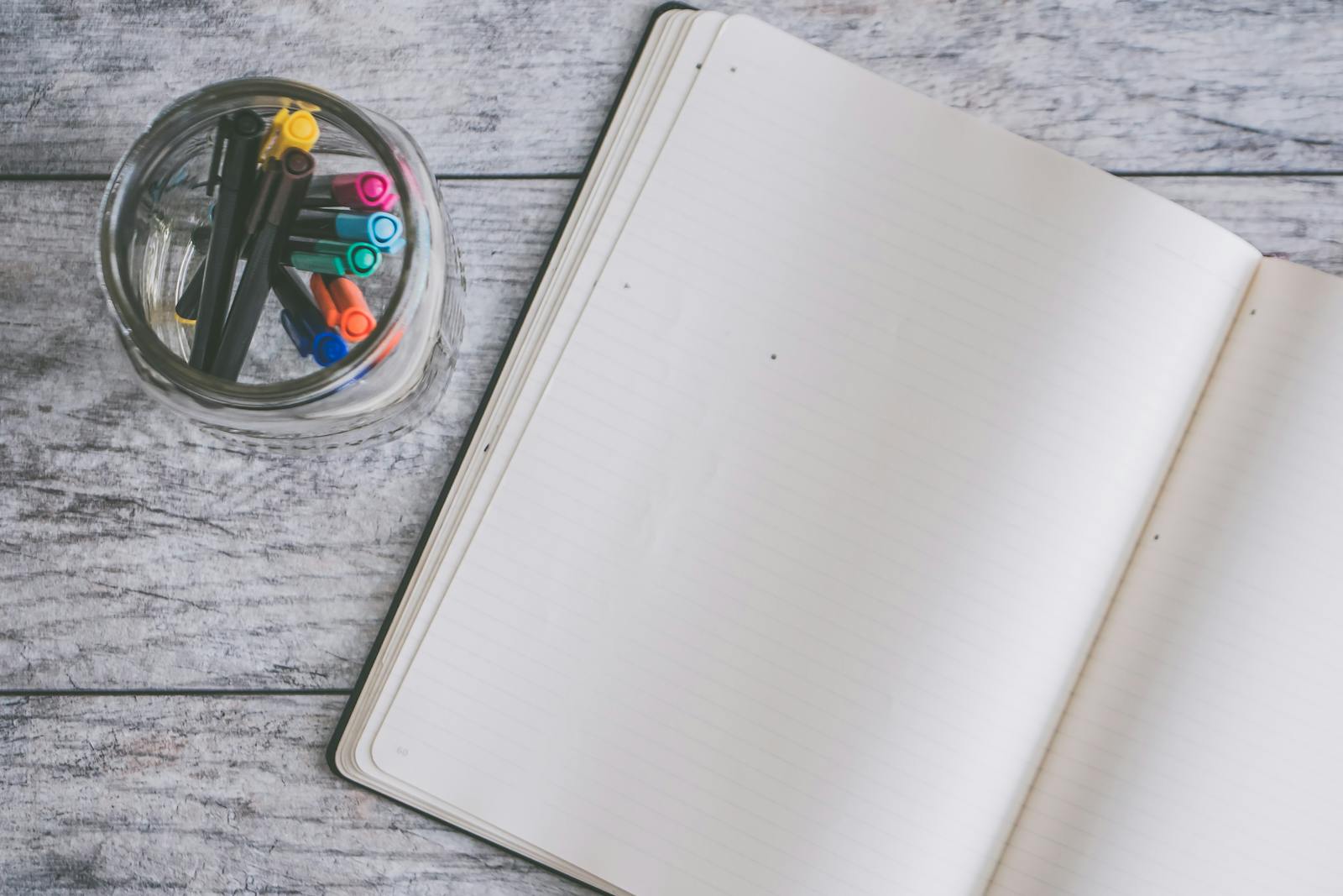 An open notebook beside a glass jar of pens, shot top-down on weathered wooden boards.