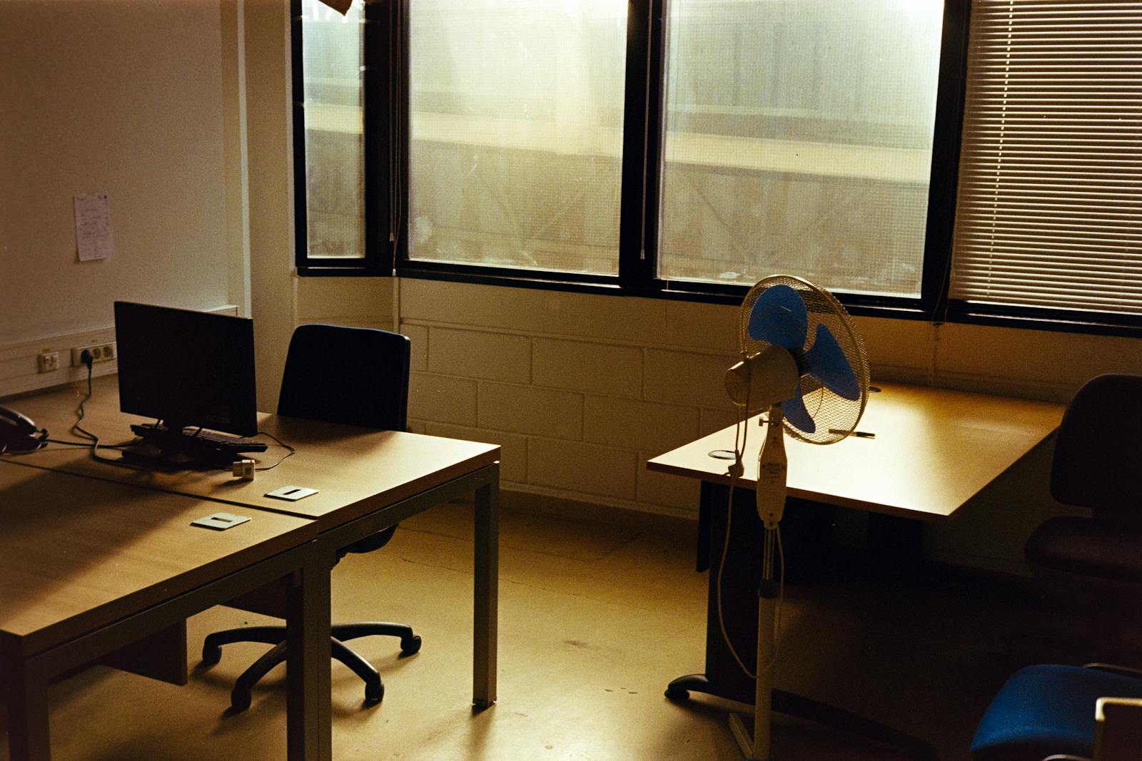 An empty office at the end of the day – desks and chairs left as they were, late-afternoon light through Venetian blinds.