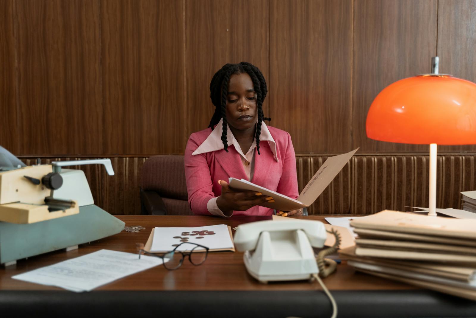 A woman in a pink blazer reads a folder at a wood-panelled desk; vintage typewriter and orange dome lamp visible.